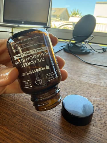 Customer holding an open brown bottle of Mini Mushroom Blend Capsules over a wooden desk.