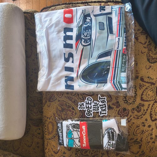 Customer's SEVEN ELEVEN TEE folded in its plastic packaging, displayed with other items on a couch.
