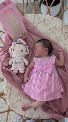 A reviewer's personalized Daisy Baby Girl doll, embroidered with the name 'Peach', lying next to a sleeping newborn baby in a bassinet.