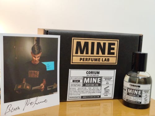 Un cliente mostra il flacone del profumo CORIUM su un tavolo, accanto alla sua confezione nera e a una foto.