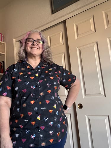Reviewer smiling while wearing the black, patterned Superman "Symbols of Hope" short sleeve shirt.