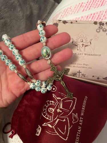 Reviewer holding the Graceful Green Floral Beads Marian Rosary, showing the crucifix over its branded pouch and card.
