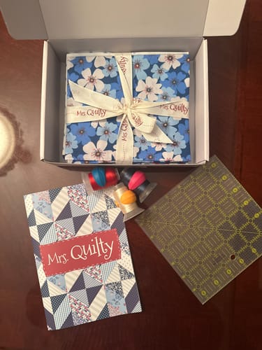Customer's Mrs Quilty Subscription Box contents including floral fabric, thread, a quilting ruler, and a branded card.