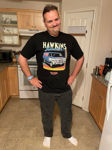 Reviewer smiling and posing in the black Hawkins Hitmaker Stranger Things t-shirt in a kitchen.