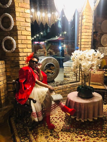 Reviewer seated indoors wearing the African Print Tulle Flared Dress, styled with a shiny red jacket and boots.