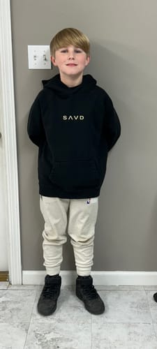 Customer's child wearing the black Youth Oversized Hoodie while standing indoors against a plain wall.