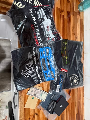 Customer's unboxing of the Ultimate Mystery Cash Box, showing t-shirts, a toy car, cash, and other accessories on a table.