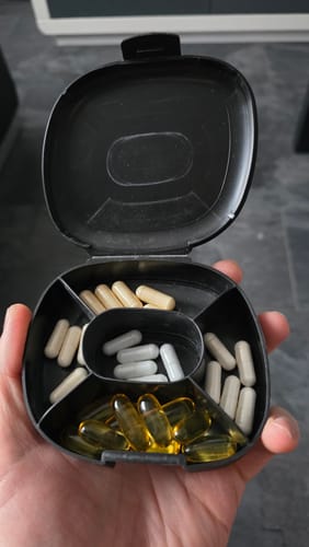 Reviewer's hand holding the open black pill box, showing its five compartments filled with various supplement capsules.