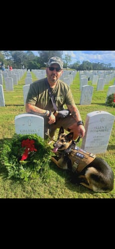 Reviewer in a green T.V.P. Elite Membership t-shirt kneeling at a gravesite with a service dog and a wreath.