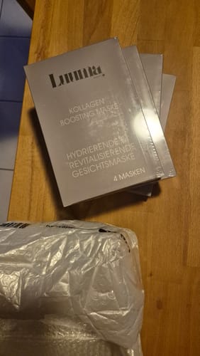 Kunde zeigt mehrere Packungen der Kollagen Boosting Maske auf einem Holztisch.