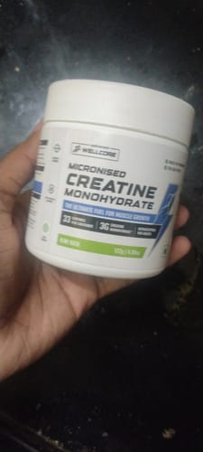 A reviewer holds up a white tub of Wellcore Micronised Creatine Monohydrate in the Kiwi Kick flavor.