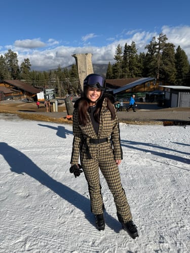 Reviewer standing on a snowy mountain in the tan houndstooth one-piece ski suit with a faux fur hood.
