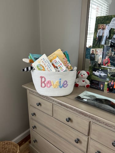 Reviewer's white Personalized Knitted Basket with "Bowie" in multicolor yarn, holding children's books and toys on a dresser.