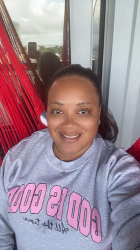 Reviewer smiling while wearing the grey 'God Is Good' sweatshirt with pink text on a balcony.
