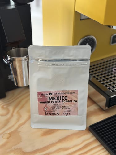 Customer's white pouch of Mexico Women Power Zongolica coffee on a counter with an espresso machine in the background.