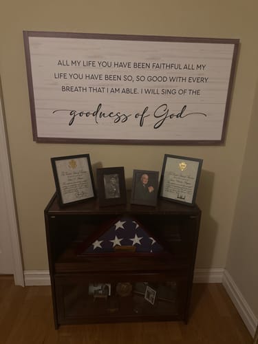 Customer's 'Goodness of God Faith Sign' on a wall above a cabinet with photos and a folded American flag.