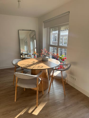 Recensent toont vier greige Nordic eetkamerstoelen die rond een ronde houten eettafel in een lichte kamer staan.