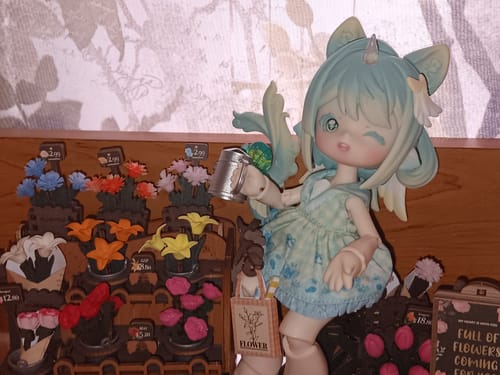 Reviewer's Antu action figure with blue-green hair, winking and holding a watering can in a miniature flower shop setting.