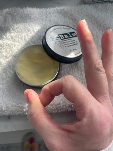 A reviewer's hand makes an okay sign over an open, round tin of F-Balm.
