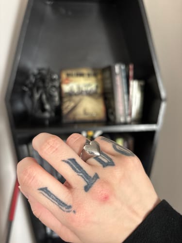 Reviewer's HEART RING on a tattooed hand, with a dark coffin-shaped shelf in the background.