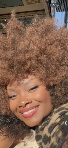 Reviewer smiling in a voluminous, light brown Janet Collection AFRO BADU wig.