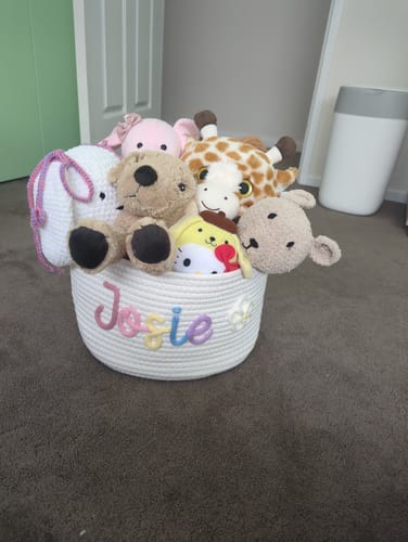 Reviewer's personalized white knitted basket with 'Josie' in multicolor yarn, filled with stuffed animals on a floor.