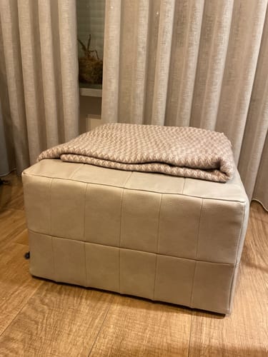 Recensent toont de naturelkleurige WOOOD Patchwork Poef met een plaid erop in een woonkamer.