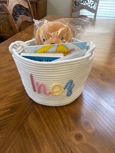 Customer's personalized Baby Knits basket with the name 'Inez' in multicolor yarn, filled with books and a stuffed puppy.