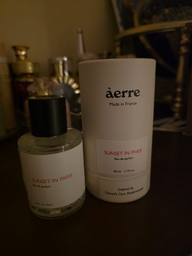 Customer's Sunset in Paris perfume bottle next to its white cylindrical packaging on a dark surface.