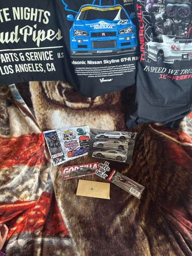 Customer's haul from the TCG90 MYSTERY CASH BOX, showing three graphic t-shirts, stickers, and an envelope.