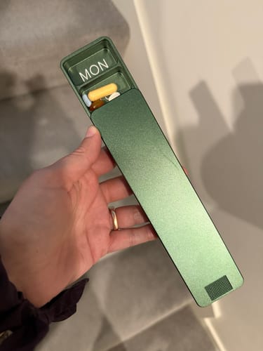 Customer holding a green metallic pill case, partially open to show the 'MON' compartment with pills inside.