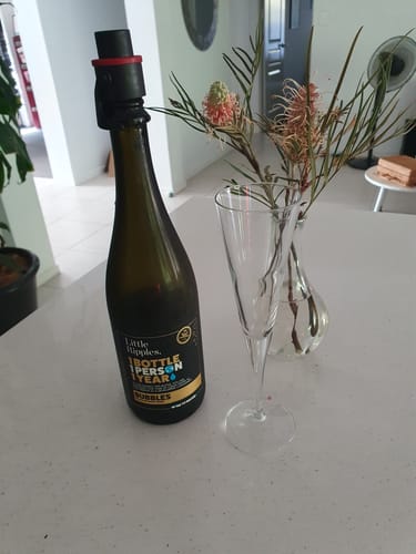 Reviewer's bottle of Little Ripples Sparkling wine with a stopper on a counter next to a tall, empty champagne flute.