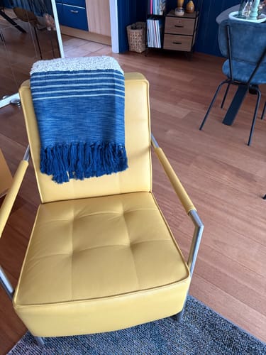 Recensent toont de blauwe Malagoon Tribal plaid, gedrapeerd over de rugleuning van een gele fauteuil.