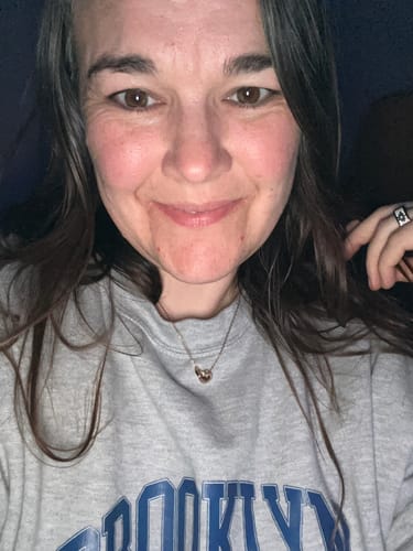 A reviewer smiling while wearing the Rose Gold Infinity Necklace with a grey sweatshirt.