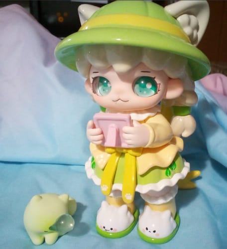 Customer's cute Mioco Seasonal Spells figurine in a green hat and yellow dress, with its small adorable cat companion.