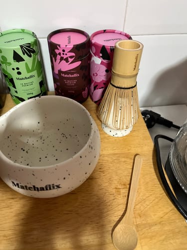 Cliente muestra el batidor chasen y la cuchara de bambú sobre una encimera de madera, junto a un bol y botes de té.