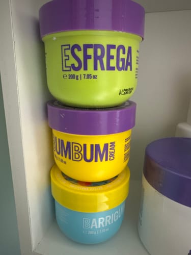 Cliente mostra os três potes coloridos do Kit Trio Cuidado Diário empilhados em uma prateleira branca.