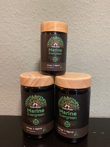 Ein Kunde zeigt drei gestapelte Dosen des Omega 3 Algenöls mit dunklen Gläsern und Holzdeckeln.
