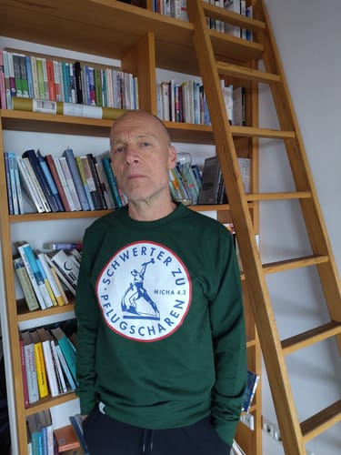 Rezensent trägt den grünen Pullover 'Schwerter zu Pflugscharen' vor einem Bücherregal.