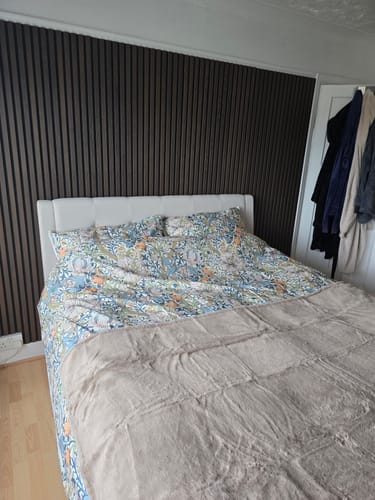 Partial view of a customer's bedroom showing dark Premium Acoustic Wall Panels installed as a feature wall behind a bed.