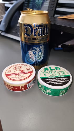 Reviewer's tin of Spicy Cinnamon Caffeine Pouches on a desk next to another tin and a drink can.