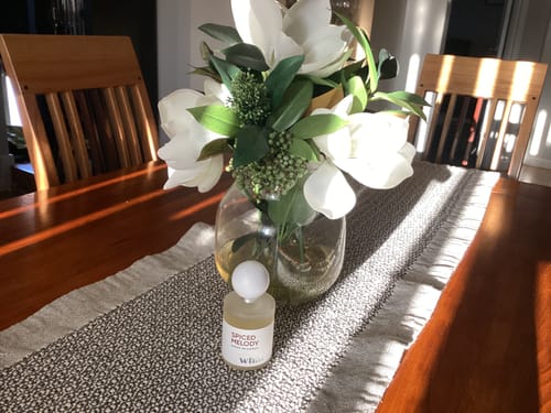 Customer's bottle of Spiced Melody on a sunlit wooden table, placed next to a vase of white flowers.