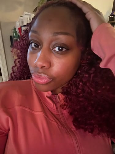 Customer wearing the red, curly Sensationnel Dream Chaser clip-in weave and a pink jacket.