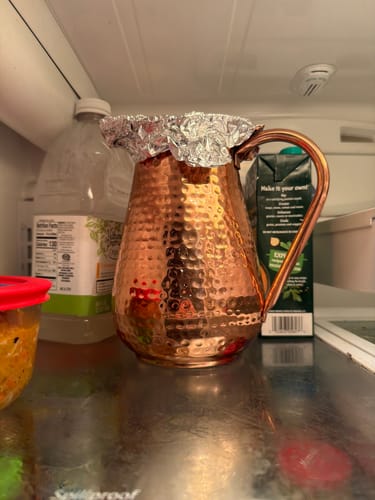 Reviewer's artisan hammered copper pitcher sitting inside a refrigerator with its top covered by foil.