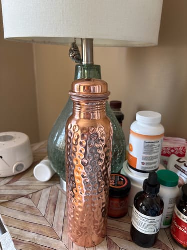 Customer's hand-hammered Pure Copper Water Bottle standing on a table next to a lamp and other bottles.