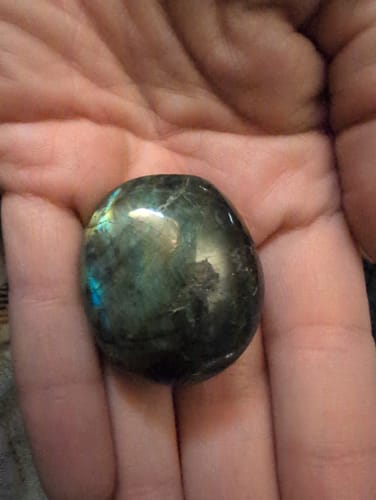 Customer's hand holding a dark, polished monthly crystal with an iridescent blue-green sheen.