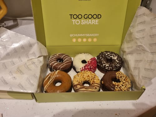 Reviewer's open box showing six assorted bronuts with chocolate, sprinkle, and nut toppings.