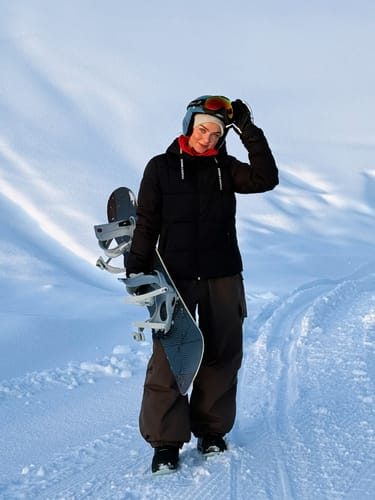 Reviewer standing in the snow holding a snowboard while wearing the baggy waterproof snow pants.