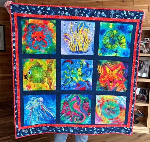 Reviewer holding a vibrant quilt with sea creatures outlined using Aurifil 40wt Mako Cotton Thread.