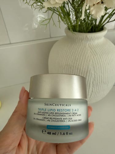 Customer's hand holding a frosted glass jar of SkinCeuticals Triple Lipid Restore cream in a bathroom setting.